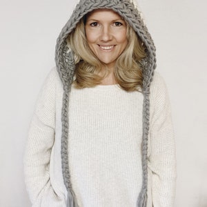 Puede incluir: Una bufanda con capucha de ganchillo blanca y gris con largos flecos. La bufanda est&aacute; hecha con hilo grueso y tiene un dise&ntilde;o trenzado.