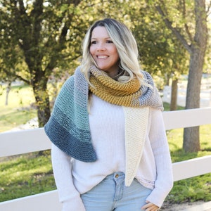 May include: A woman wearing a white sweater and blue jeans is standing in front of a white fence. She is wearing a multi-colored knitted scarf with a yellow stripe.