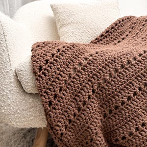 May include: A brown crochet blanket draped over a cream armchair. The blanket has an open, textured weave, and the chair has a rounded back and wooden legs. A cream pillow rests behind the blanket.