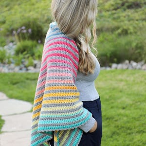 May include: A woman wearing a striped crochet shawl with a variety of colors, including pink, blue, yellow, and gray. The shawl is crocheted in a simple stitch and has a scalloped edge.