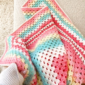 以下が含まれることがあります： ピンク、イエロー、グリーン、ホワイトの色のグラニー・スクエア模様のクロシェ編みベビーブランケット。ブランケットは半分に折り畳まれています。