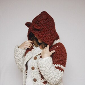 May include: A crocheted cardigan with a hood in white and burgundy. The cardigan has a button closure and a geometric pattern.