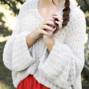 May include: A woman wearing a cream-colored, fluffy sweater with wide sleeves and red pants. The sweater has a V-neck and a textured knit pattern. The woman's hair is in a braid.