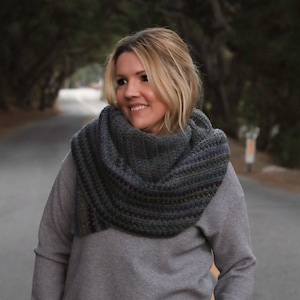 May include: A woman wearing a gray sweater and a gray and green striped scarf. She is standing on a paved road with trees in the background.