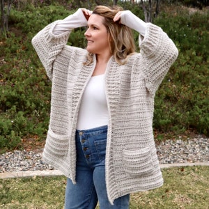 Op de afbeelding: Een beige gehaakte cardigan met een shawlkraag en twee zakken. De cardigan is open aan de voorkant en heeft een losse pasvorm.
