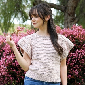 May include: A woman wearing a light pink crocheted top with ruffled sleeves and blue jeans. She is standing in front of a bush of pink flowers.