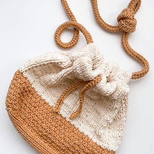 Może przedstawiać: Tkana torba ze sznurkiem do ściągania z długim paskiem. Torba wykonana jest z dwóch kolorów przędzy: jasnego beżu i brązu. Torba jest zebrana u góry i ma sznurek do ściągania.