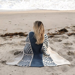 May include: A person sitting on a sandy beach wearing a blue and white crocheted blanket. The blanket has a geometric pattern and is draped over the person's shoulders.