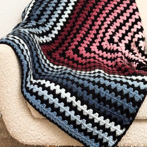 Może przedstawiać: Ręcznie robiony koc zrobiony na szydełku w wzór granny square. Koc ma gradient kolorów, w tym czarny, niebieski, biały i odcienie różu i czerwieni. Jest narzucony na beżowy fotel, prezentując swoją fakturę i wzór.