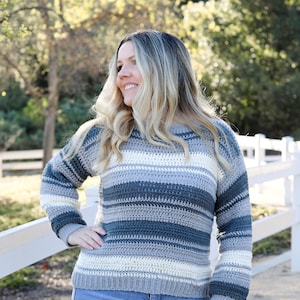 May include: A striped crocheted sweater in shades of gray, cream, and teal. The sweater has a relaxed fit and long sleeves.