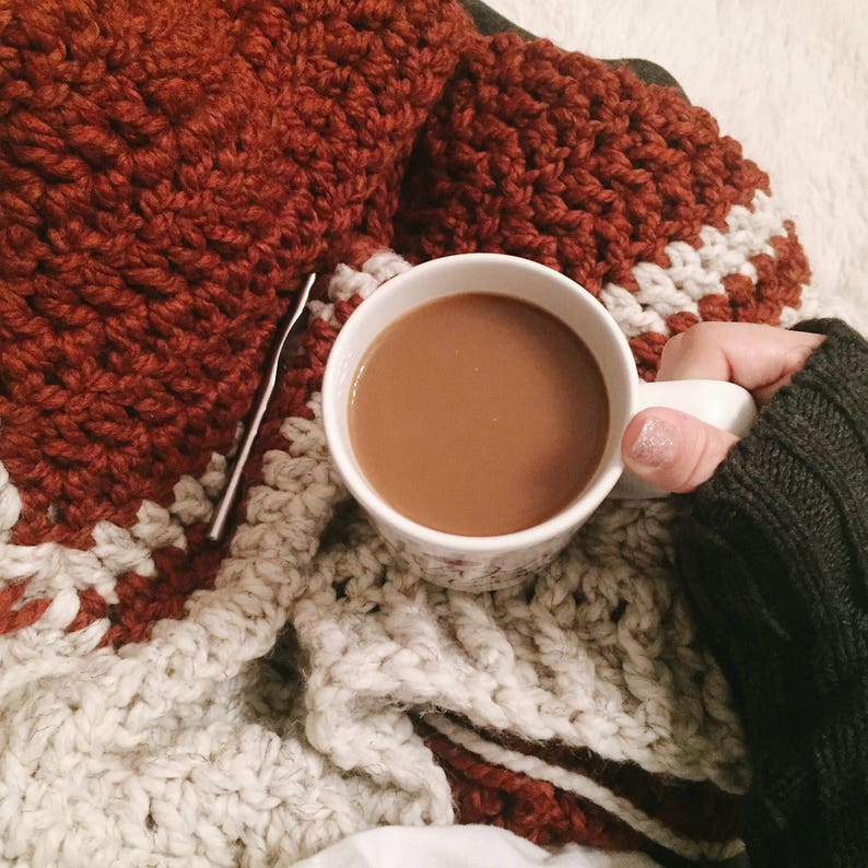 May include: A white ceramic mug filled with a brown beverage, held by a hand wearing a dark grey sweater. The mug is resting on a crocheted blanket with brown and white stripes.