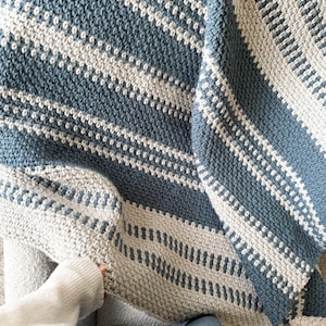 May include: A blue and white striped crocheted blanket with a textured pattern. The blanket is folded and draped over a chair.