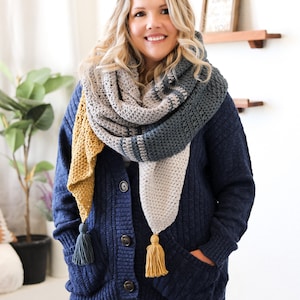 Może przedstawiać: Kobieta w niebieskim kardiganie z dzianym szalem w odcieniach szarego, niebieskiego i żółtego. Szal ma frędzle na końcach.
