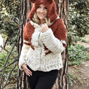 May include: A crocheted hooded sweater with a fox design. The sweater is white and brown with a brown hood and fox ears. The sweater is being worn by a person standing in front of a tree.