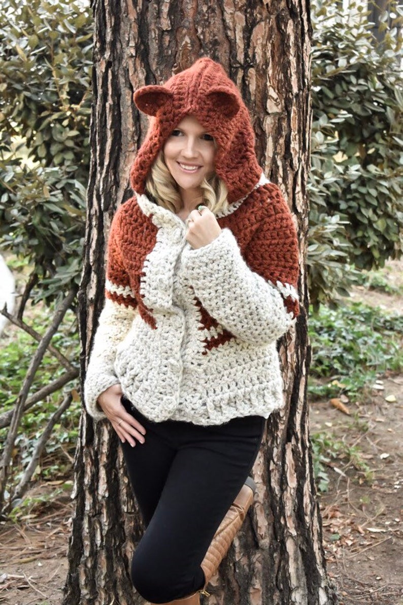 May include: A crocheted hooded sweater with a fox design. The sweater is white and brown with a brown hood and fox ears. The sweater is being worn by a person standing in front of a tree.