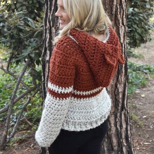 May include: A crocheted hooded sweater with a rust coloured body and white sleeves. The sweater has a white crocheted trim around the hood and sleeves.