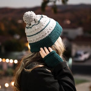 May include: A green and white knitted beanie with a pom pom on top. The beanie is being worn by a person with long brown hair. The person is also wearing a green knitted fingerless glove.