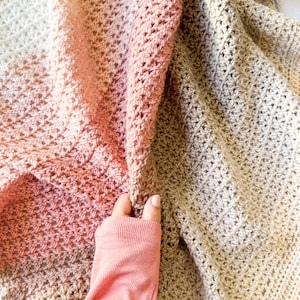 May include: A crocheted throw blanket with a gradient of peach, cream, and beige colors. The blanket is folded in half and a hand is holding the corner of the blanket.