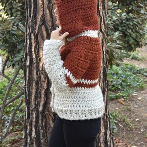 May include: A brown and white crocheted cardigan with a hood and ear flaps. The cardigan has a button closure and a white trim around the edges.