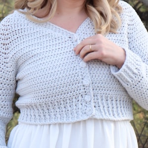 May include: A light grey crocheted cardigan with long sleeves and a button fastening. The cardigan is cropped and has a slightly loose fit.