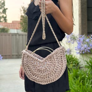 May include: A beige crochet handbag with a gold-tone metal handle and chain strap. The bag has a rounded shape and is held by a person wearing a black dress. The background includes greenery and a building.