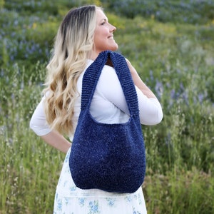 Puede incluir: Una mujer con una blusa blanca y una falda floral lleva un bolso tote de punto azul sobre el hombro. El bolso tiene un estilo informal y holgado y está hecho con un hilo texturizado.