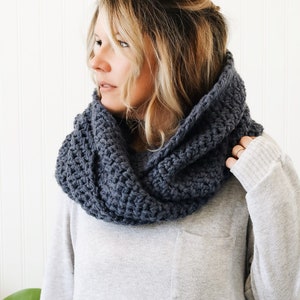 May include: A gray crocheted infinity scarf worn around the neck of a person with long blonde hair.
