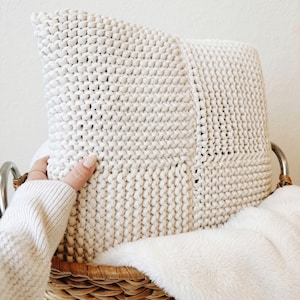 May include: A white knitted throw pillow in a wicker basket with a white faux fur blanket.