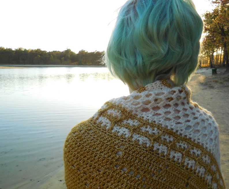 May include: A person with teal blue hair wearing a crocheted shawl with a white and gold pattern. The shawl is draped over the person's shoulders and back. The person is standing in front of a body of water with trees in the background.