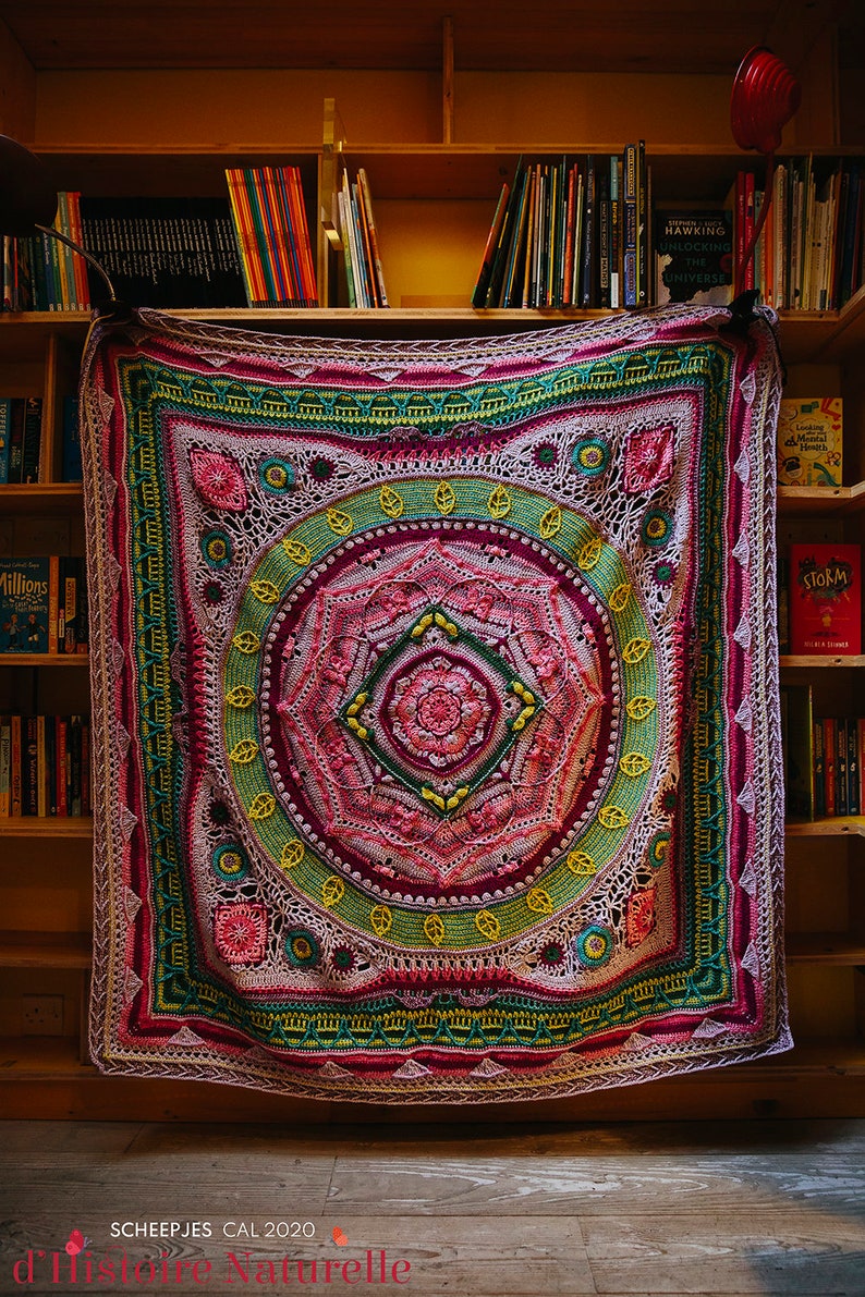 May include: A large, colorful crochet blanket with a mandala design in shades of pink, green, and yellow. The blanket is hanging on a bookshelf with books visible on the shelves. The text "SCHEEPJES CAL 2020" is visible at the bottom of the image.
