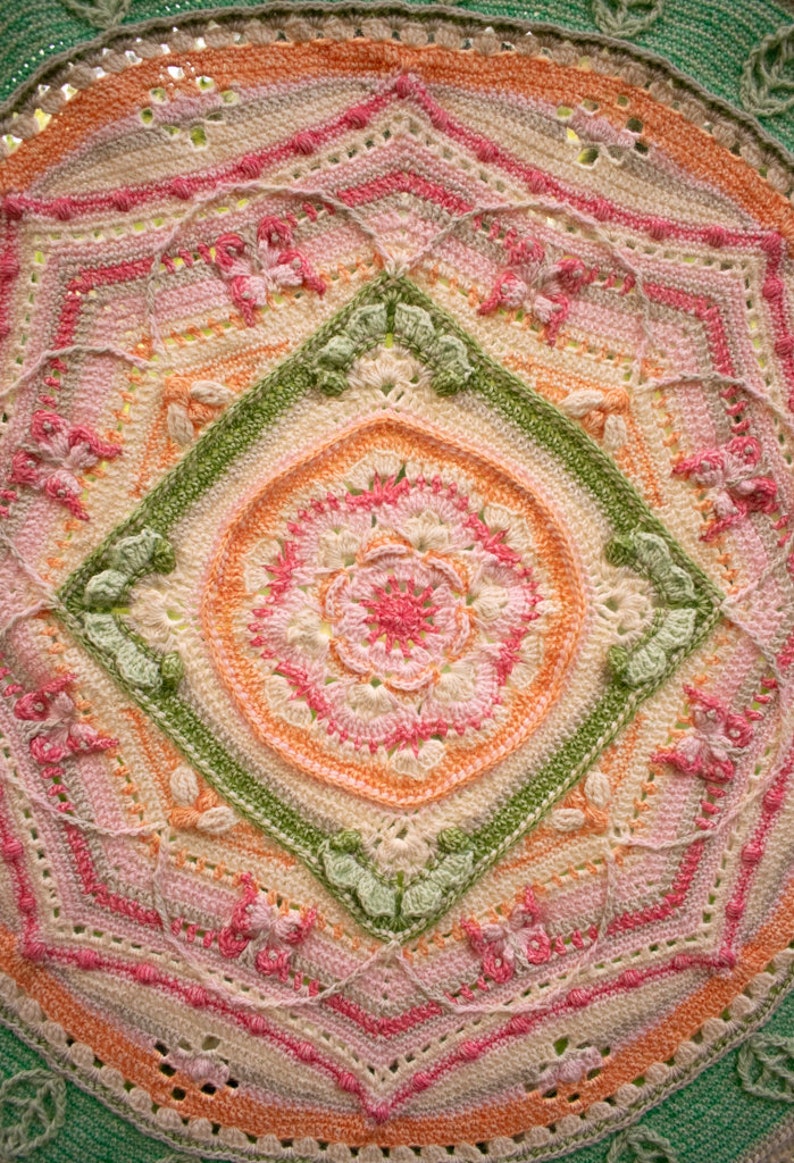 May include: A crocheted mandala with a central flower design in shades of pink, orange, and green. The mandala is surrounded by a border of leaves and flowers.