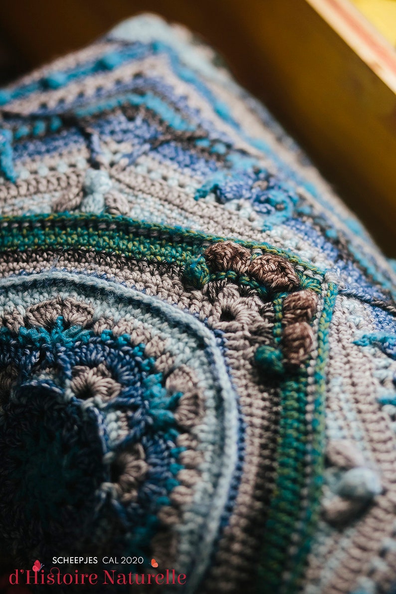 May include: Close-up of a crocheted blanket with a blue, gray, and green color scheme. The blanket has a textured pattern and is made with a variety of stitches. The text "SCHEEPJES CAL 2020 d'Histoire Naturelle" is visible in the bottom right corner.