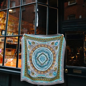 May include: A large, colorful crochet blanket with a mandala design in shades of blue, green, yellow, and brown. The blanket is being held up in front of a storefront with the text "Scheepjes Cal 2020 d'Histoire Naturelle" at the bottom.