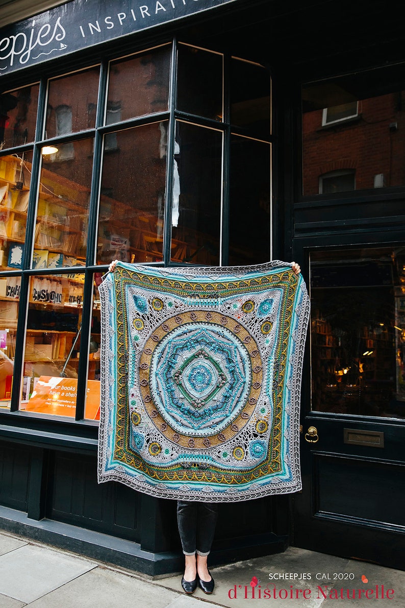 May include: A large, colorful crochet blanket with a mandala design in shades of blue, green, yellow, and brown. The blanket is being held up in front of a storefront with the text "Scheepjes Cal 2020 d'Histoire Naturelle" at the bottom.