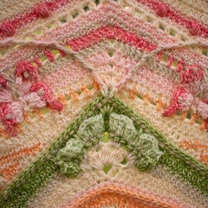 May include: Close-up of a crocheted blanket with a pink, green, and white color scheme. The blanket features a repeating pattern of flowers and butterflies.