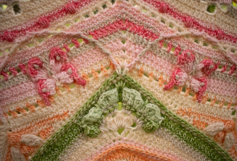 May include: Close-up of a crocheted blanket with a pink, green, and white color scheme. The blanket features a repeating pattern of flowers and butterflies.