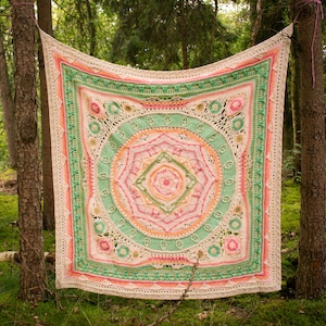 以下が含まれることがあります： ピンク、グリーン、ホワイトの色の円形のマンダラ模様が施された、かぎ針編みブランケット。ブランケットは森の中に生えている2本の木の間にかけられています。
