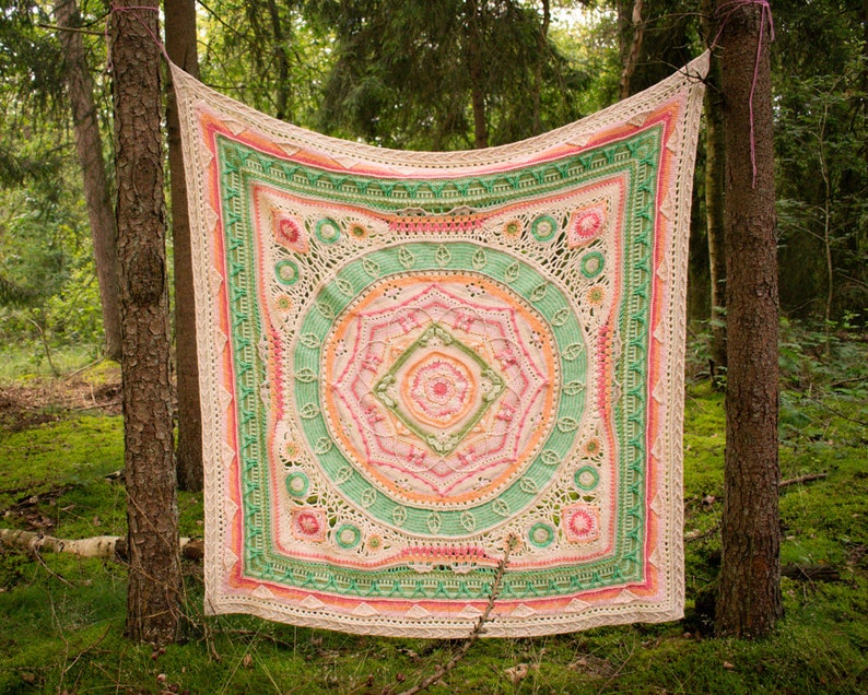 May include: A crocheted blanket with a circular mandala design in shades of pink, green, and white. The blanket is hanging between two trees in a forest setting.