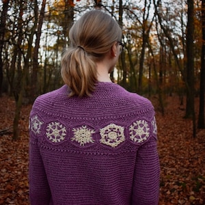 Op de afbeelding: Een paarse gehaakte cardigan met een wit sneeuwvlokkenontwerp op de rug. De cardigan wordt gedragen door een persoon die in een bos staat.