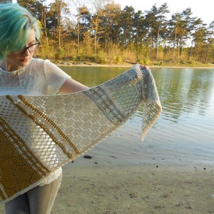 May include: A woman with teal blue hair is holding a crocheted shawl with a white, beige, and gold pattern. The shawl is draped over her arm and she is standing in front of a lake.