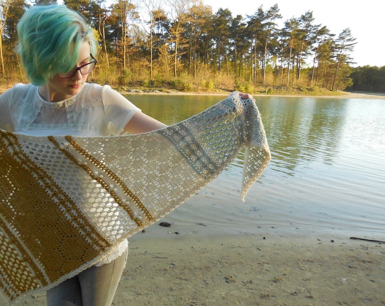 May include: A woman with teal blue hair is holding a crocheted shawl with a white, beige, and gold pattern. The shawl is draped over her arm and she is standing in front of a lake.