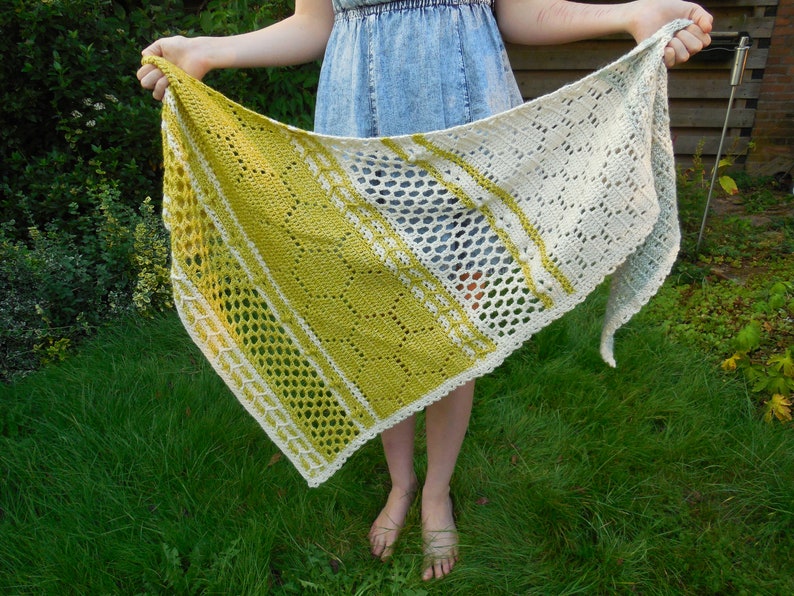 May include: A crocheted shawl with a white and green color gradient. The shawl has a lacy pattern and is held by a person in a garden.