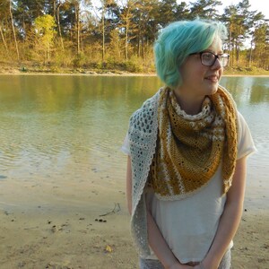 May include: A woman with teal blue hair wearing a white shirt and a crocheted shawl with a light blue and mustard yellow color pattern. The shawl is draped around her neck and shoulders. She is standing in front of a body of water with trees in the background.