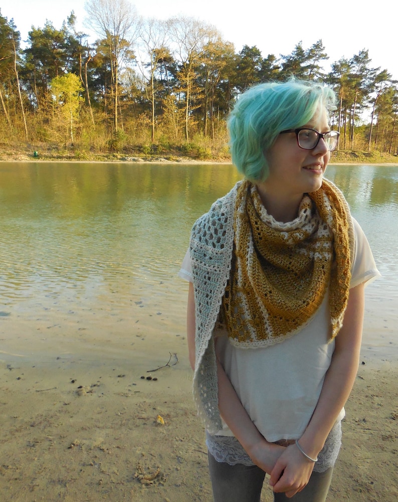 May include: A woman with teal blue hair wearing a white shirt and a crocheted shawl with a light blue and mustard yellow color pattern. The shawl is draped around her neck and shoulders. She is standing in front of a body of water with trees in the background.