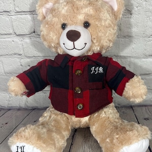 May include: A plush teddy bear with tan fur, wearing a red and black plaid shirt with the initials "J.I.B." embroidered on the pocket. The bear's foot pads have the text "I'll always with you" and "Grandpa".