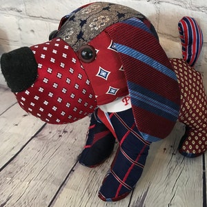 May include: A handmade dog-shaped plush toy crafted from repurposed neckties. The dog features a black felt nose, large button eyes, and is pieced together with various patterned fabrics in shades of red, blue, and patterned designs.