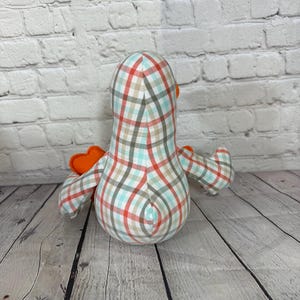 Pu&ograve; includere: Un peluche a forma di uccello in tessuto fatto a mano, visto da dietro, con un motivo a quadri rossi, turchese e marrone. Ha ali e becco arancioni. Il peluche &egrave; su una superficie di legno.