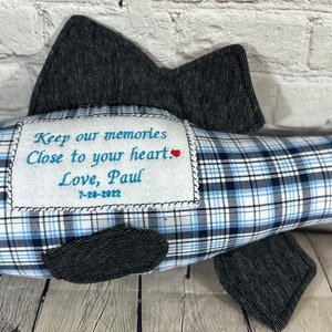 Op de afbeelding: Een blauw en wit geruit visvormig kussen met een grijze stoffen achterkant. Het kussen heeft een klein geborduurd hartje en de tekst "Keep our memories close to your heart. Love, Paul 7-28-2022".