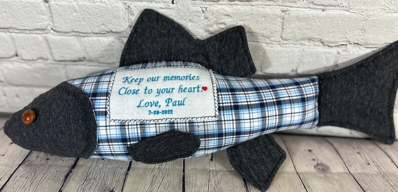Op de afbeelding: Een blauw en wit geruit visvormig kussen met een grijze stoffen achterkant. Het kussen heeft een klein geborduurd hartje en de tekst "Keep our memories close to your heart. Love, Paul 7-28-2022".