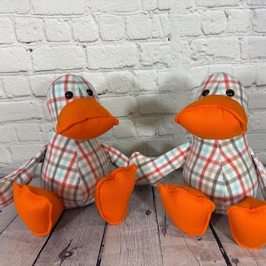 Pu&ograve; includere: Due giocattoli a forma di anatra di peluche con becchi e piedi arancioni. Le anatre hanno corpi a fantasia a quadri rossi, blu e marroni. I giocattoli sono su una superficie di legno davanti a un muro di mattoni bianchi.